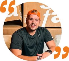 Rubens Stuque, fundador do Sofá Na Caixa, sentado em um sofá de braços abertos, com uma camisa preta e um boné laranja em primeiro plano, e ao fundo, estão caixas de cores marrom do produto Sofá Na Caixa